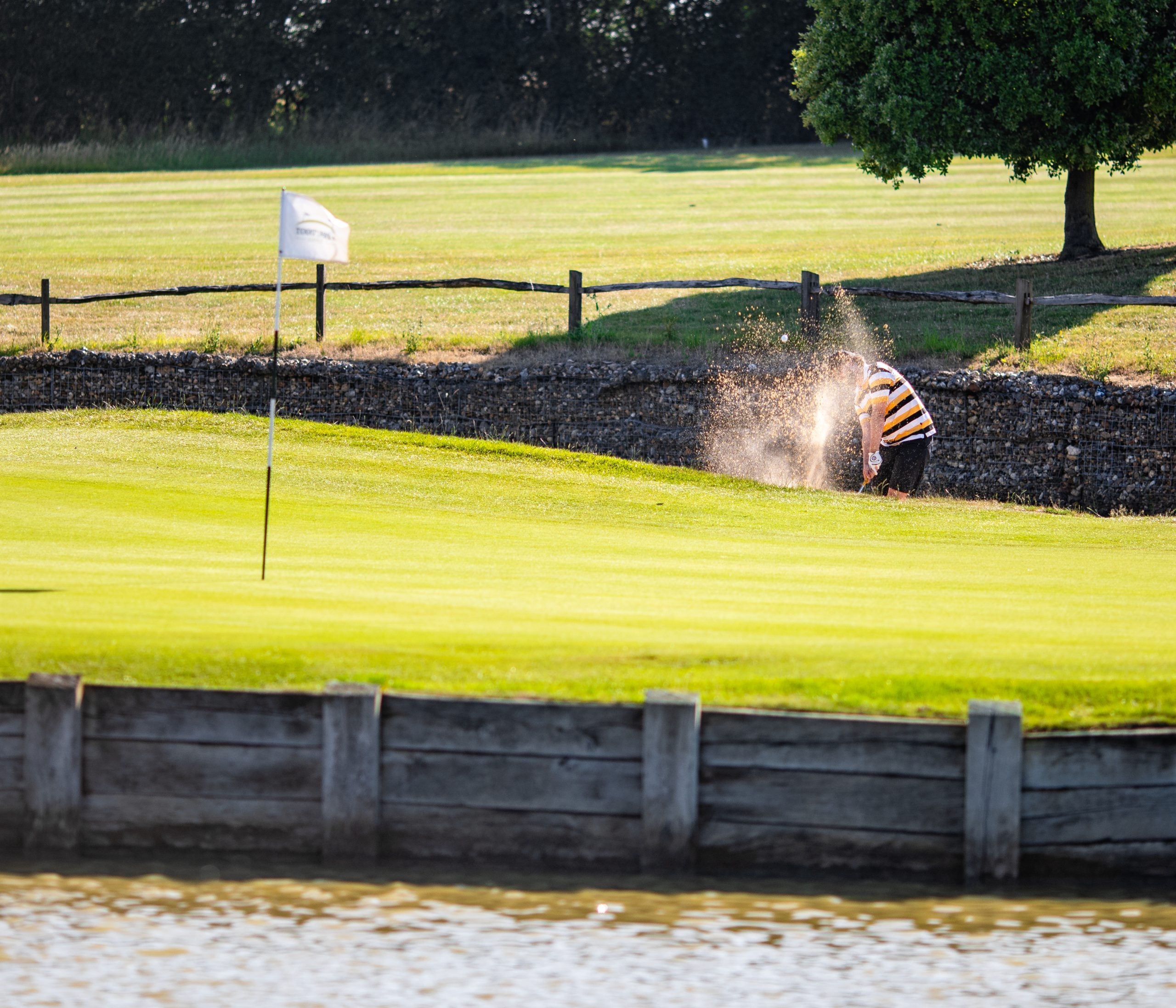 Golfer playing out of a bunker near green next to water feature on a sunny day on Toot Hill golf course